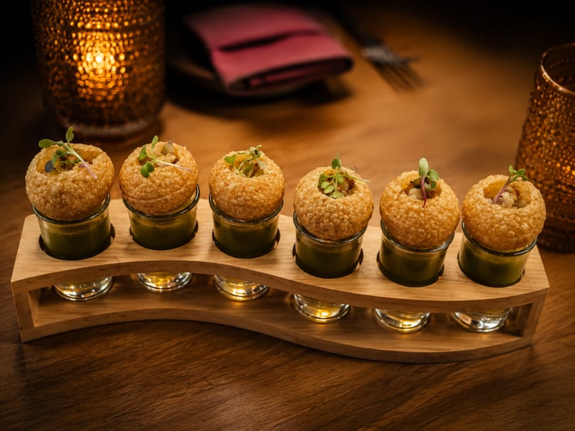 Six pani puri appetizers with green sauce in shot glasses on a wooden tray, with candlelight.