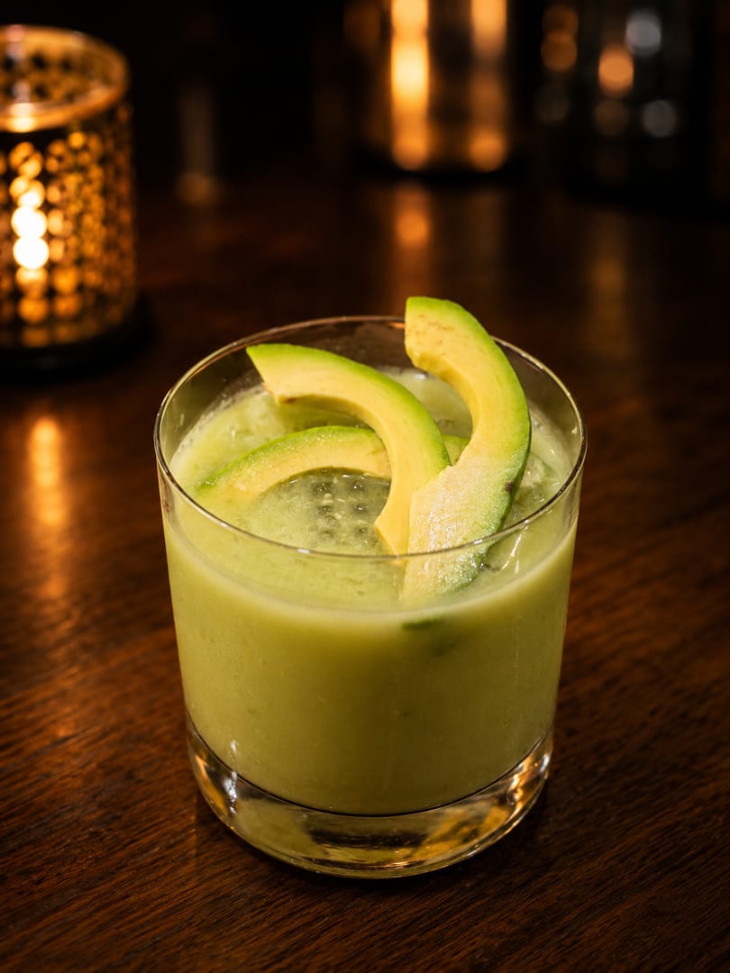 Avocado cocktail garnished with avocado slices on a dark wooden table with blurred candlelight.