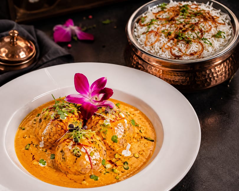 Three kofta balls in orange gravy, garnished with microgreens and an orchid, with rice.