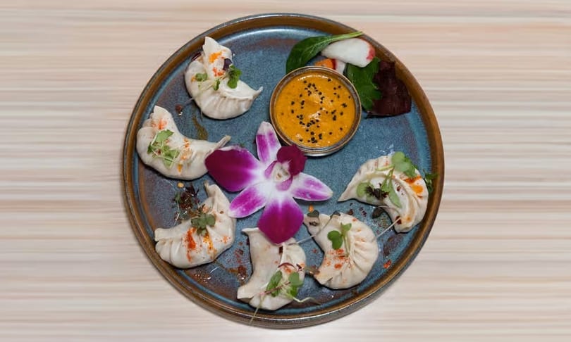 A blue plate with six dumplings, a small bowl of orange sauce, and a purple orchid.