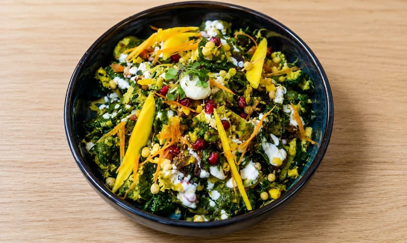 A vibrant green salad with mango, pomegranate, and white dressing in a dark bowl.