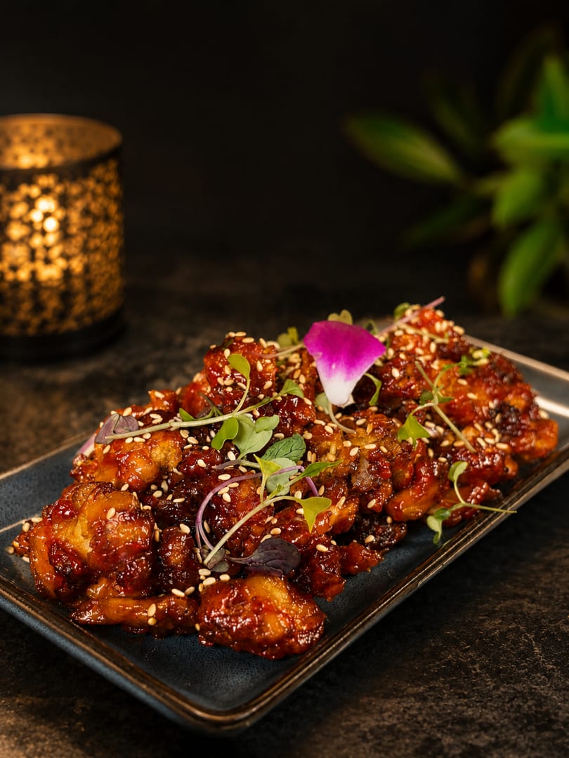 Sweet and sour chicken with sesame seeds and microgreens on a dark plate, with a candle in the background.